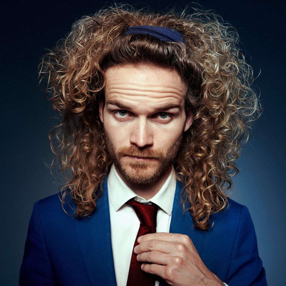 Foto van Teun met een enorme bos krullend haar. Hij draagt een blauw colbert, rode stropdas en blauwe haarband en staat voor een blauwe achtergrond.