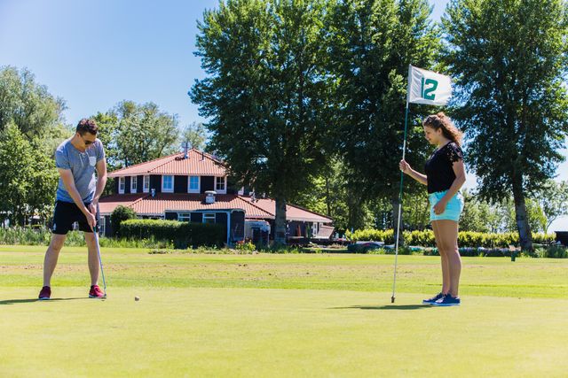 Man en vrouw spelen Pitch & Putt op het zonnige grasveld bij Buitenplaats Kameryck in Woerden, met clubhuis op de achtergrond