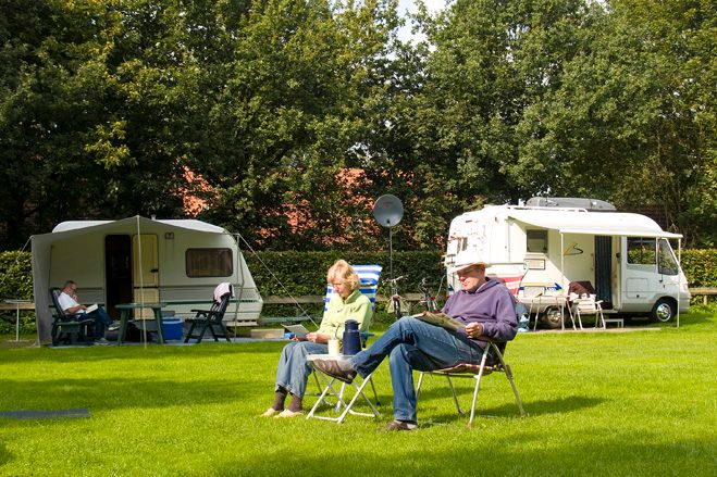 Foto van een groen grasveld met aan de rand bomen, waaronder 2 caravans staan en 2 gasten op hun campingstoeltjes op de voorgrond.