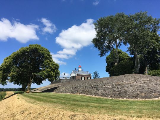 Een kleine heuvel waar bomen omheen staan. Op de heuvel staat een klein huisje. De lucht is helder en blauw. In Kraggenburg, de Noordoostpolder.