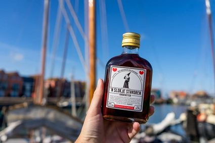 iemand houd een flesje Slokje van Stavoren vast met de haven van Stavoren op de achtergrond