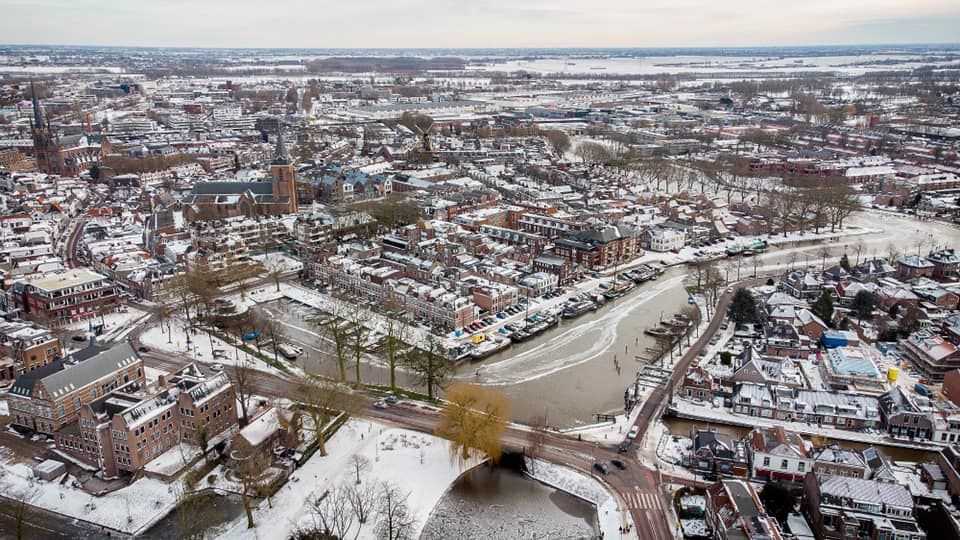 Een indrukwekkende winterse luchtfoto van Woerden, waar de stad onder een zachte sneeuwdeken ligt en de kronkelende grachten deels bevroren zijn. In de haven trekken schaatsers hun sporen over het ijs, terwijl de historische binnenstad en omliggende wijken een betoverende, rustige wintersfeer uitstralen. Een prachtig overzicht van Woerden in haar meest idyllische seizoen.