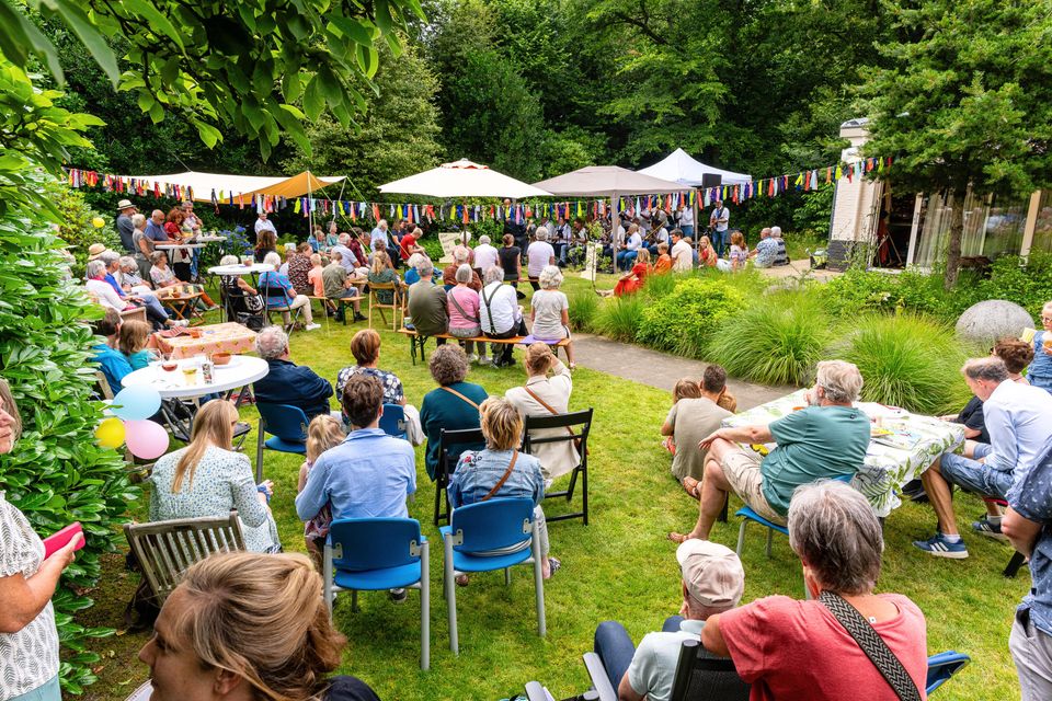 Publiek kijkt naar een muzikaal optreden in een kleurrijk versierde tuin tijdens Gluren bij de Buren in Gemeente Woerden, met stoelen, parasols en een festivalsfeer.