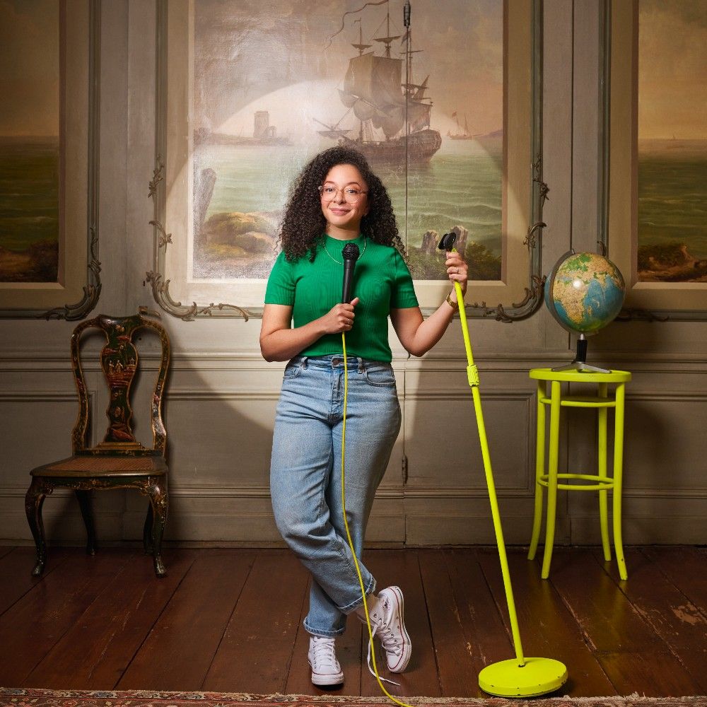 Wina Ricardo met lange donkere krullen in een groen truitje staat voor een klassiek schilderij en een felgroene kruk met wereldbol daarop. Ze heeft ook een felgroene microfoonstandaard vast en microfoon in de hand.