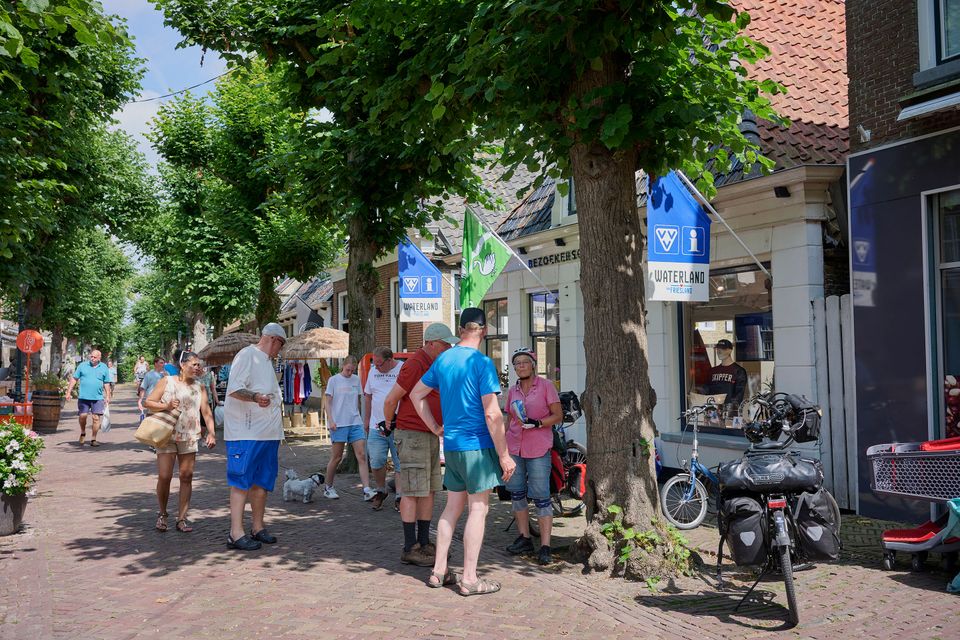Meerdere zomers geklede mensen staan voor het VVV bezoekerscentrum in Langweer. Er hangen blauwe vlaggen uit.