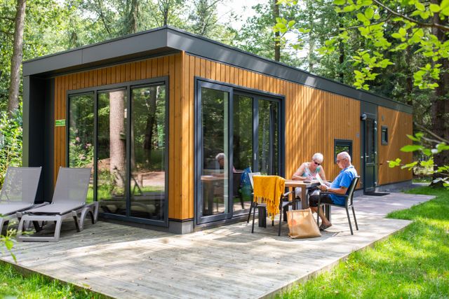 Modern houten vakantieverblijf Doornveld met terras bij RCN het Grote Bos. Twee gasten zitten buiten voor de bungalow in de bosrijke omgeving.