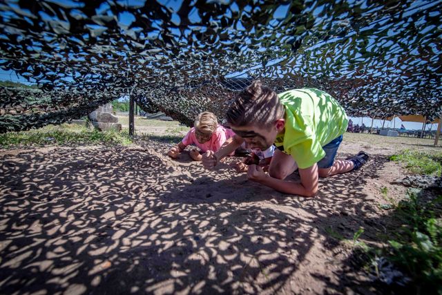 Kinderen kruipen onder een camouflagenet tijdens een actief buitenparcours bij het Nationaal Militair Museum in Soest. De foto laat een speelse en avontuurlijke activiteit voor kinderen zien.