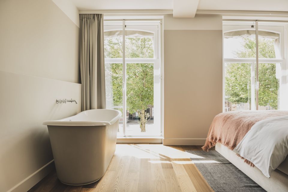 Lichte hotelkamer met grote ramen. Links staat een vrijstaand bad en rechts staat het bed, opgemaakt met een roze sprei.
