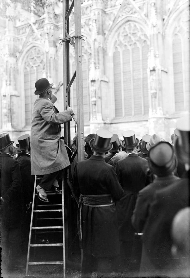 0028924 Burgemeester van Lanschot, zittend op een trapleer en steunend op een elektriciteitsmast geeft uitleg over de kerk juni 1935