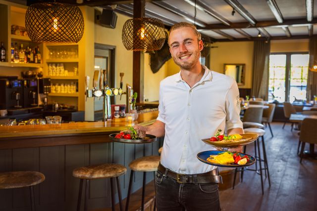 Glimlachende ober in wit overhemd serveert drie borden met kleurrijke gerechten in Bistro en Bar De Dijketelg in Oudewater, met op de achtergrond de sfeervolle houten bar en warme verlichting.