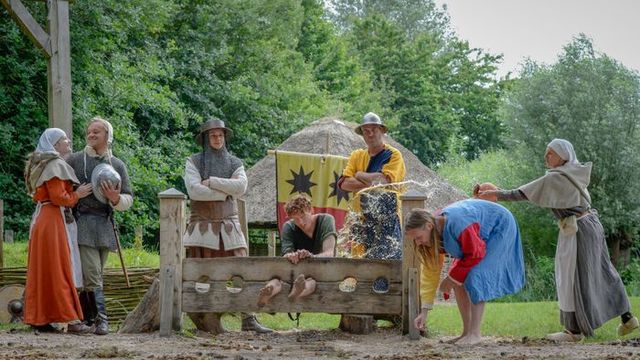 Groep mensen in middeleeuwse kleding voert een ludieke strafscène uit met een schandpaal in Archeon, als onderdeel van een interactieve geschiedenisbeleving in het Groene Hart.