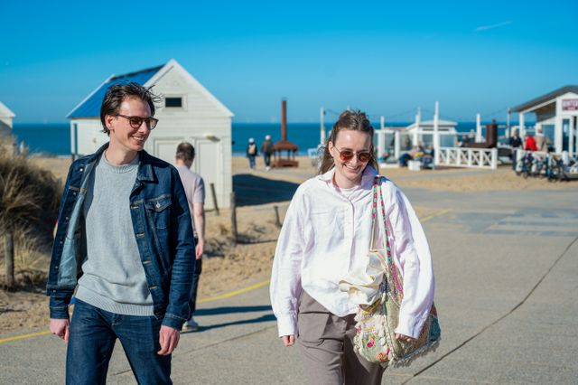 Jong koppel loopt lachend in Katwijk aan Zee, met de achtergrond een strandpaviljoen en de zee.