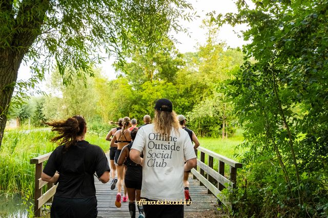 Natuurgebied in Woerden, Groene Hart, social run van de Offline Outside Social Club met hardlopers die over een houten brug door een groen park en langs het water rennen tijdens een gezamenlijke hardloopactiviteit.