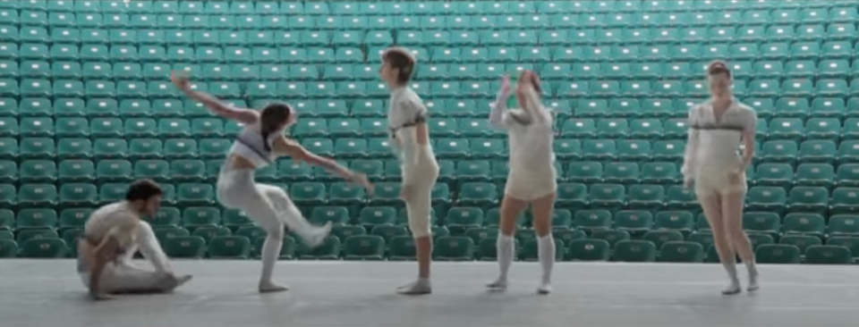 Vijf dansers in witte kostuums voeren een moderne choreografie uit op een leeg podium, met groene stadionstoelen als achtergrond.