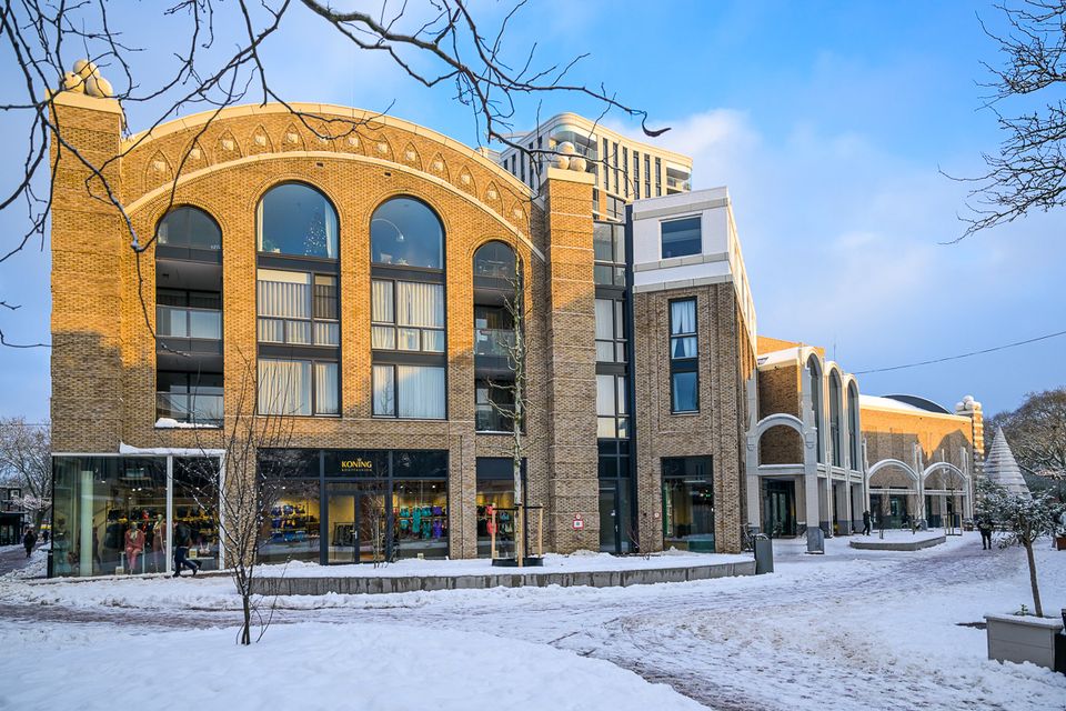 Groot bakstenen gebouw met boogvormige ramen en winkels op de begane grond. Voor het pand ligt een besneeuwd plein met wandelpaden en bomen zonder bladeren. Op de achtergrond zijn moderne woongebouwen zichtbaar onder een blauwe winterlucht.