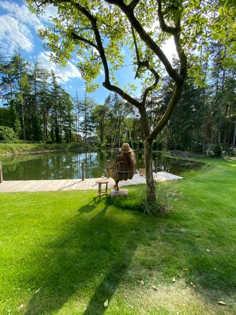 Grasveld met boom met hangstoel waarin een gaste zit. Aan een vlonder bij de vijver.