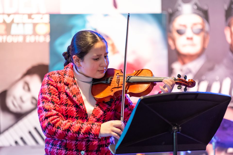Meisje speelt viool vanaf een lessenaar tijdens een optreden op een podium, geconcentreerd kijkend naar de bladmuziek, met posters van musici op de achtergrond.