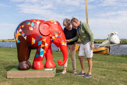 Twee mensen bewonderen een van de kleurrijk beschilderde olifanten van de Elephant Parade in Sloten en Gaasterland met op de achtergrond een boot.