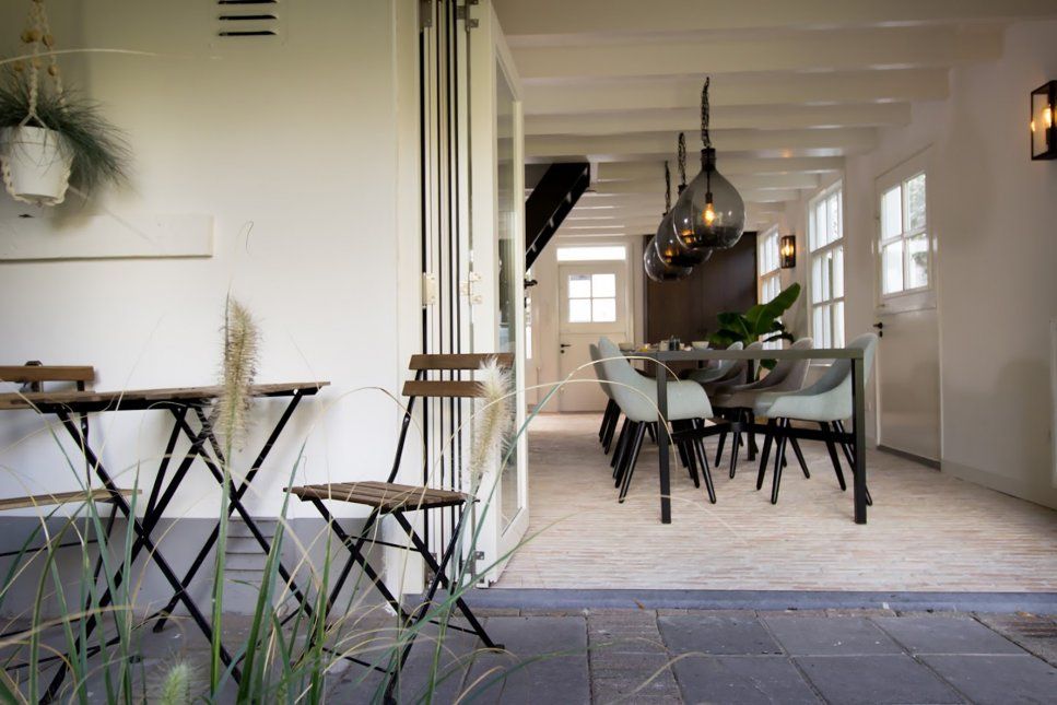 Doorkijkje naar eettafel met kuipstoeltjes binnen, openslaande deuren en een bistro-zitje op de voorgrond buiten tegen een witte gevel.