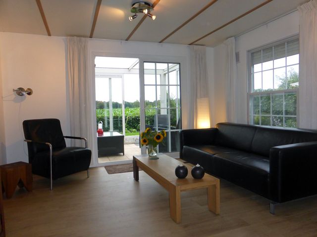 Woonkamer met een zwart leren bankstel en fauteuil rond een salontafel. Openslaande deuren naar de serre, met uitzicht op het maisveld.