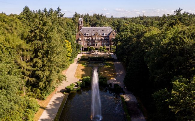 Dronefoto van Kasteel Hooge Vuursche. Met oprijlaan, omgeven door bos en een fontein op de voorgrond.