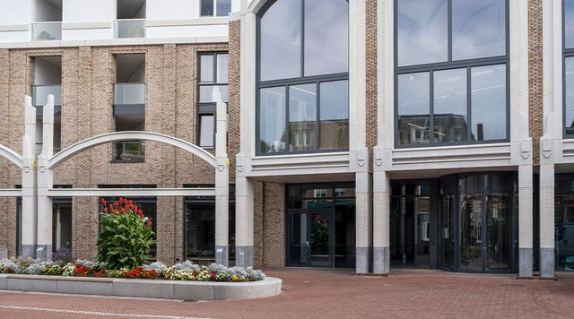 Vooraanzicht van een modern gebouw met bakstenen gevel, grote ramen en een glazen draaideur. Voor het gebouw ligt een betegeld plein met een ronde plantenbak met bloemen en groen.