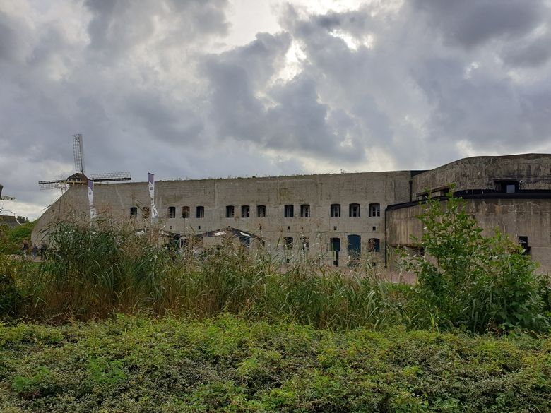 Een betonnen fort tegen een zwaar bewolkte lucht. Op de voorgrond rietkragen.