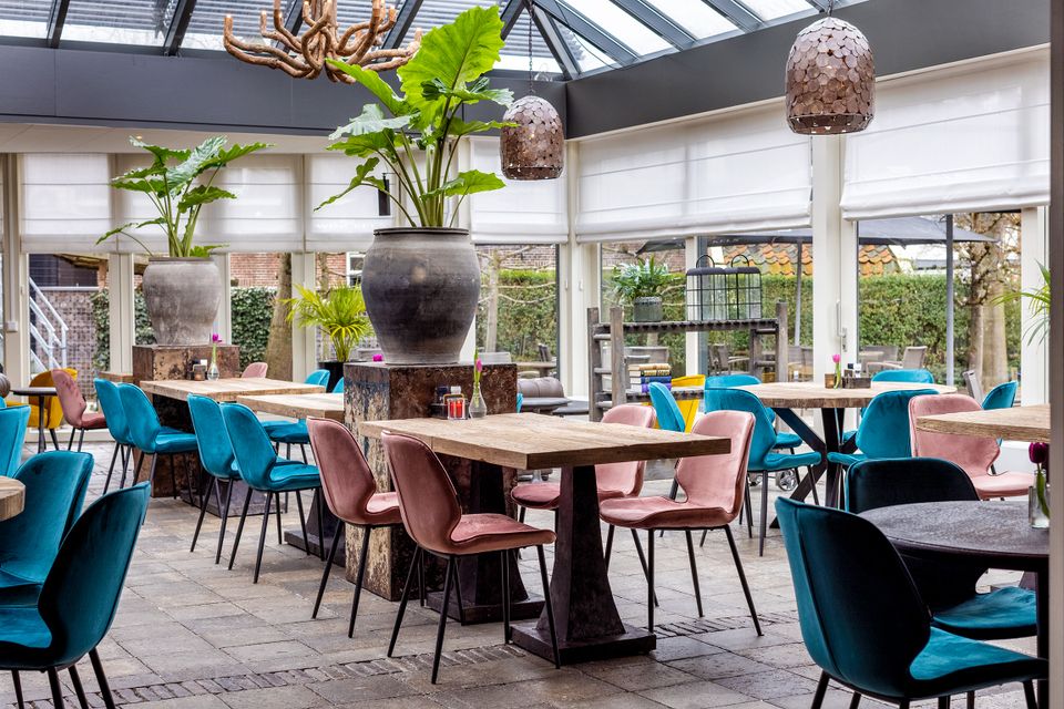 Stijlvol interieur van het restaurant van Hotel en Grand Café Broeck in Oudewater met houten tafels, kleurrijke stoelen en veel groen, gelegen in een lichte serre met uitzicht op de tuin.