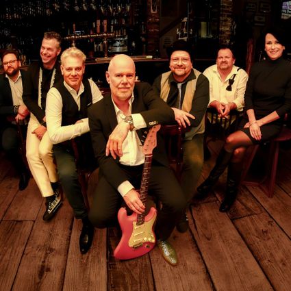 Foto van de bandleden in zwart-witte kleding op een houten vloer voor een bar. Het middelste bandlid heeft een rode gitaar vast.