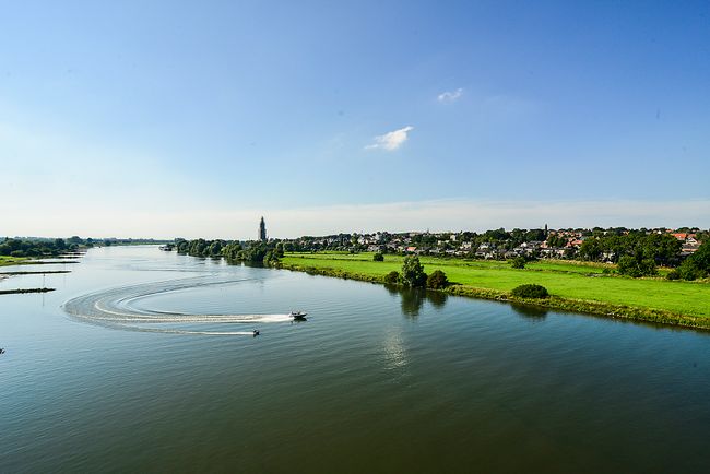 Een boot vaart over de Nederrijn. Op de achtergrond zie je groen gas met de skyline van Rhenen en de Cuneratoren.