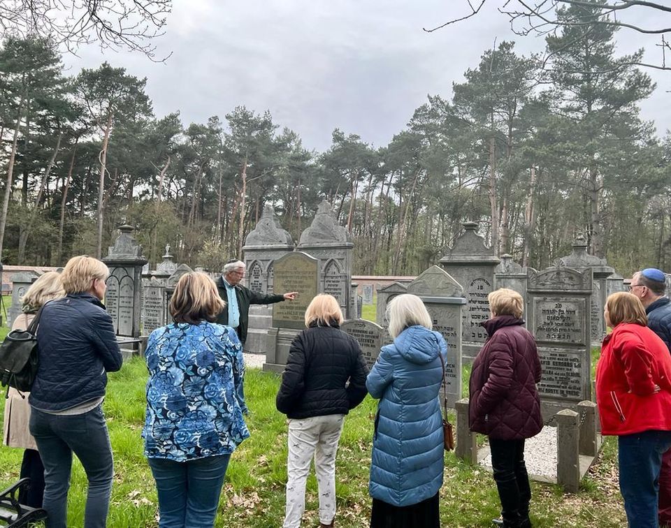 Rondleiding op de joodse begraafplaats. Een gids geeft uitleg aan een groep personen over de grafstenen. De graafplaats ligt in een groene omgeving met bomen op de achtergrond.