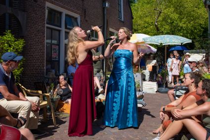 Cultureel evenement Gluren bij de Buren in de Hogewalstraat, Woerden, Groene Hart, met twee zangeressen die optreden voor publiek in een zonnige stadstuin tijdens een kleinschalig muziekfestival.