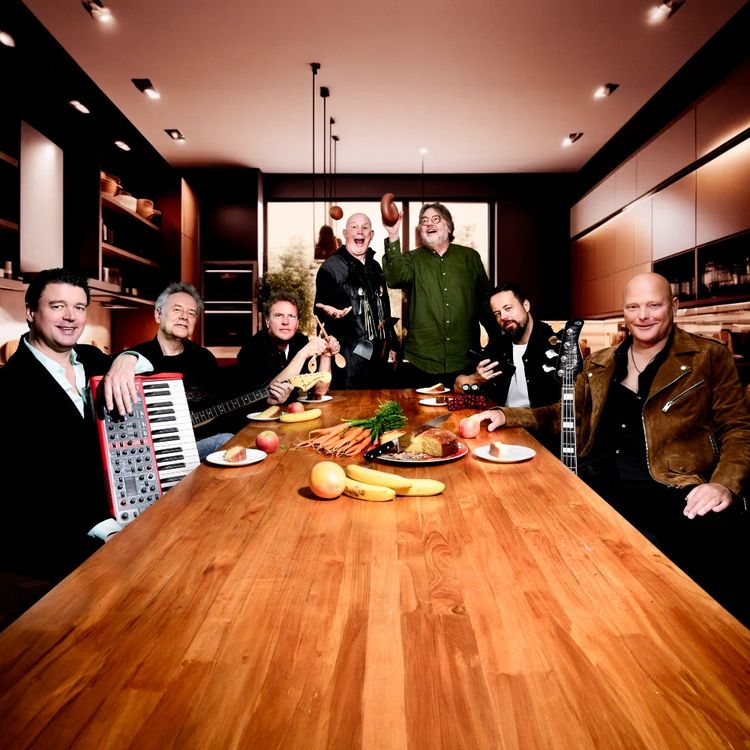 Foto van een lange houten tafel gevuld met groenten en fruit. Er om heen zitten de bandleden met hun muziekintrumenten en daarachter staan Henk Westbroek en kok Pierre Wind.