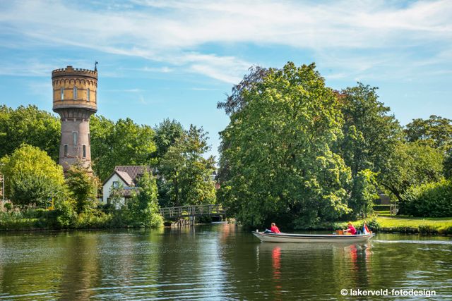 Sloep met twee personen vaart over het water in Woerden, met op de achtergrond de historische watertoren en groene bomen in zomerse sfeer.