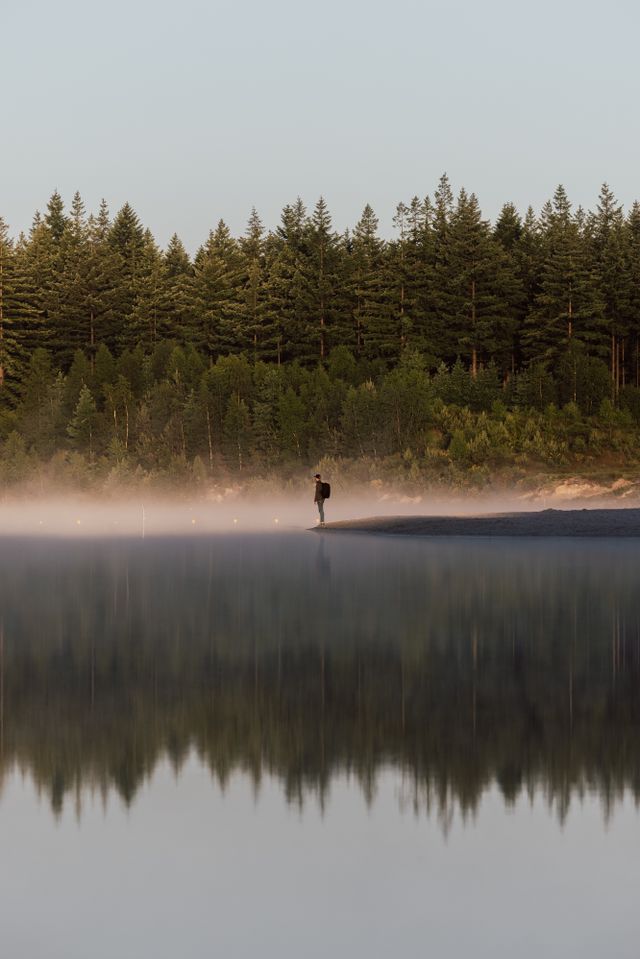 Een man staat in zijn eentje bij een zwemplas met het bos op de achtergrond.