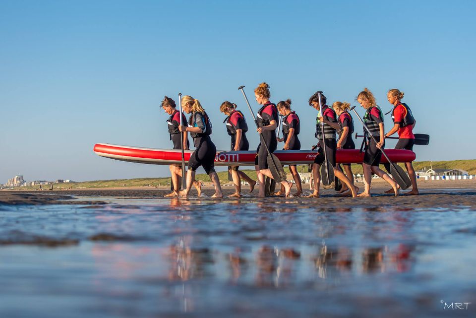 Afbeeldingen van een groep strandgangers die een megasup richting zee tillen in Katwijk.