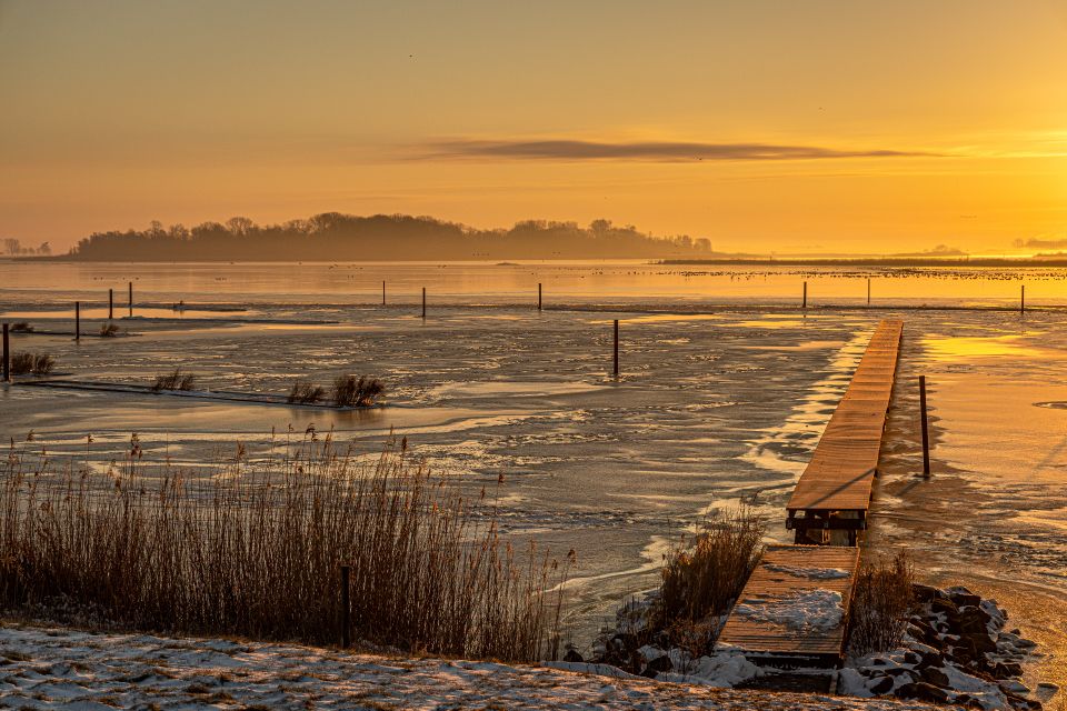 Een dijk met daarachter een pier over het water. Op de achtergrond zie je de ondergaande zon in Kraggenburg in de Noordoostpolder.