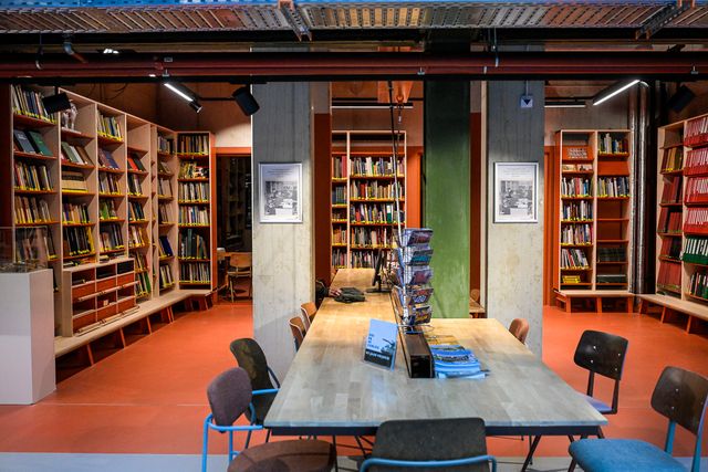 Interieur van een bibliotheek met hoge boekenkasten, een lange houten leestafel met stoelen en tijdschriften in het midden, en industriële verlichting aan het plafond.