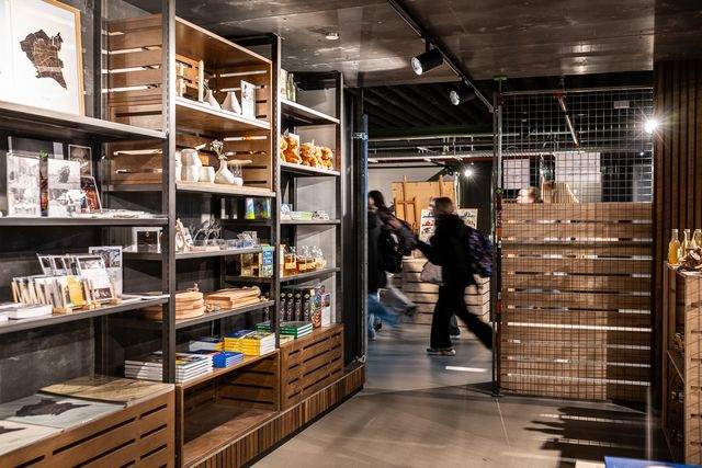Winkelruimte met open stellingkasten vol souvenirs, boeken en cadeaus, terwijl bezoekers langs de schappen lopen in een museum- of bibliotheekwinkel met industriële inrichting.