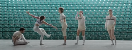 Vijf dansers in witte kostuums voeren een moderne choreografie uit op een leeg podium, met groene stadionstoelen als achtergrond.