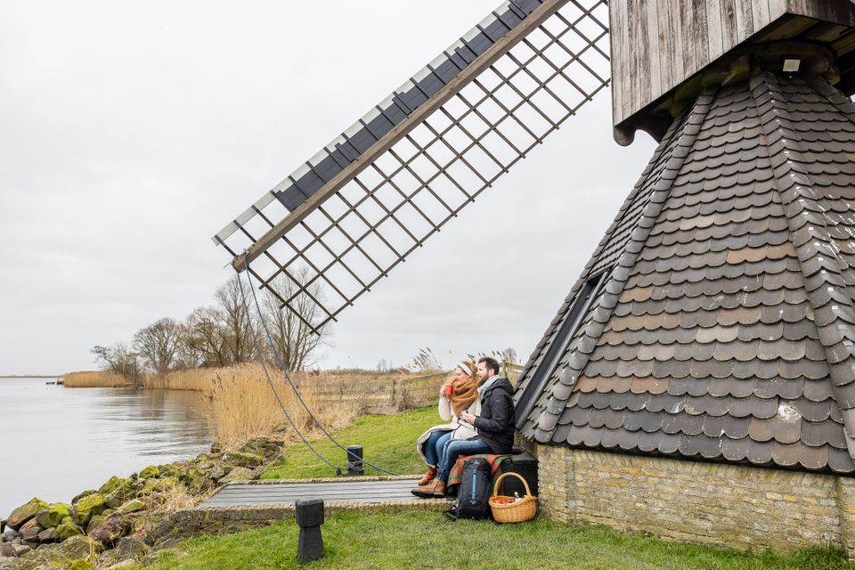 Een stel zit op een bankje tegen een pittoresk molentje, ieder een winterjas aan kan je aan de omgeving zien dat het najaar is.