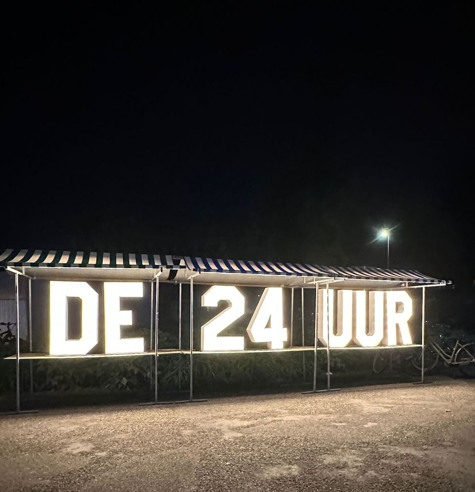 Verlicht logo van de 24 Uur van Woerden met grote lichtgevende letters ‘DE 24 UUR’, opgesteld langs een weg in het donker, onderdeel van het evenement 24 Uur van Woerden.