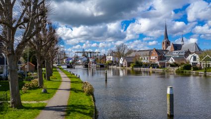 Dorpsgezicht aan een Nederlandse vaart met een wandelpad langs het water, groene oevers met bomen, woonhuizen en een kerk met hoge torenspits op de achtergrond onder een lucht met stapelwolken.