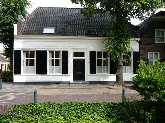 Monumentale witte voorgevel met donkere voordeur en luiken langs de ramen. Gelegen aan de doorgaande weg met veel groene beplanting.
