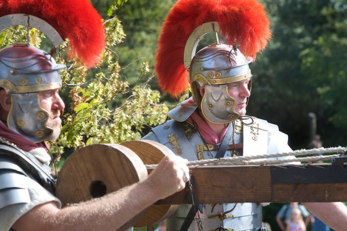 Twee Romeinse soldaten bedienen een ballista tijdens een re-enactment in Archeon, gekleed in traditioneel harnas met rood gevederde helmen.