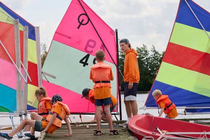 Kinderen in zwemvesten op de steiger met instructeur en zeilbootjes.