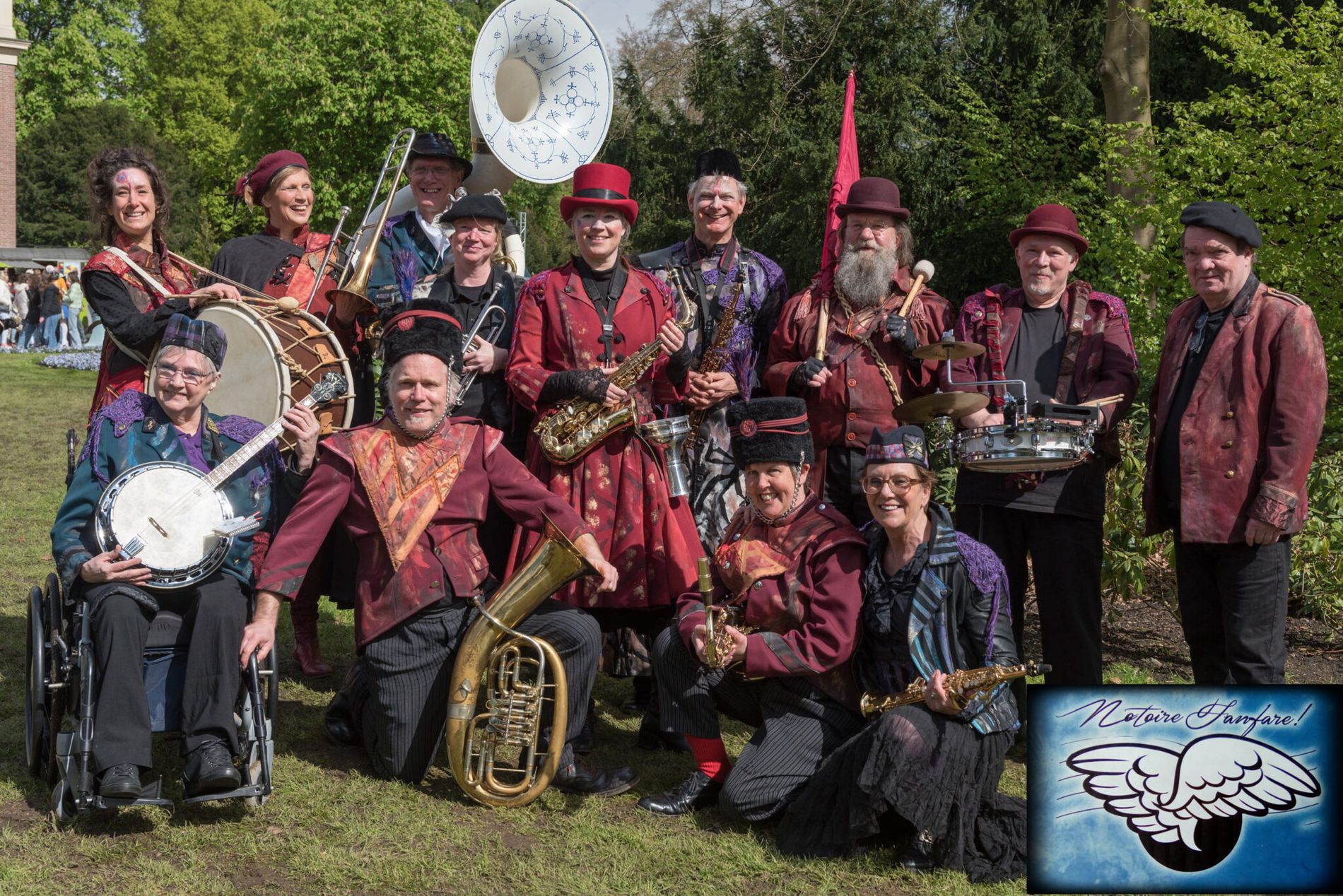 Foto van dames en heren van de fanfare met paarse en donkerrode kleding en diverse muziekinstrumenten. Ze staan in de openlucht in een bosrijke omgeving.