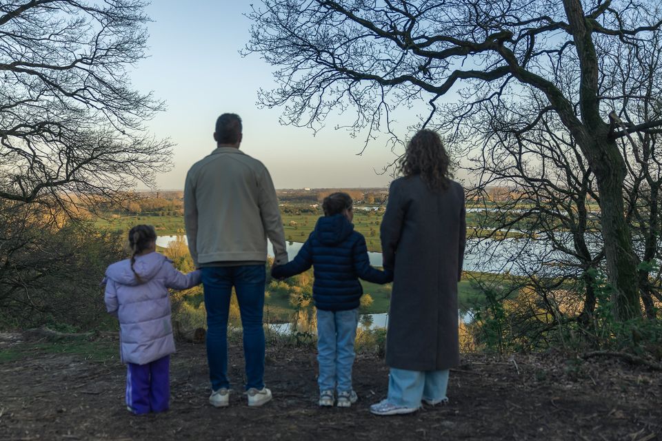 Een gezin staat hand in hand op de Grebbeberg en kijkt uit over de uiterwaarden bij Rhenen. Kale boomtakken omlijsten het weidse landschap met water, weilanden en bomen.