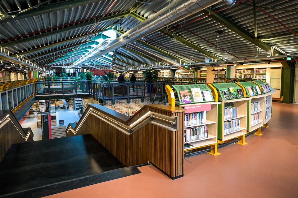 Bovenverdieping van een moderne bibliotheek met zicht op beneden: een trap met leuning, boekenkasten langs de rand, industriële plafondconstructie en bezoekers die over de vide lopen.
