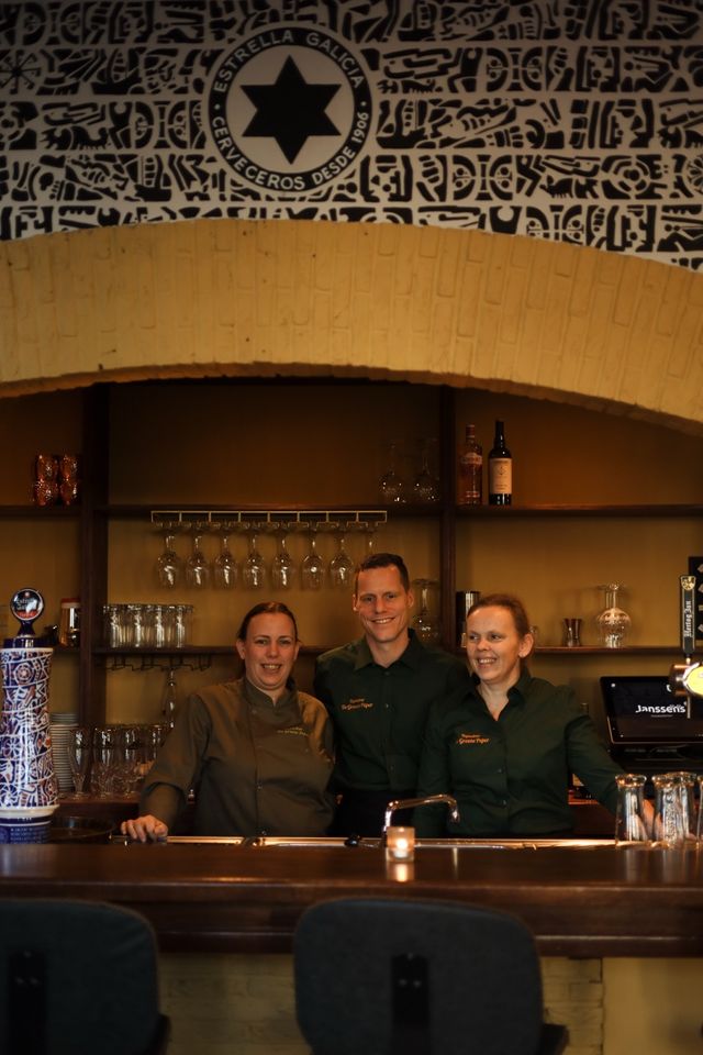 Drie medewerkers staan achter de bar in een restaurant en kijken glimlachend richting de camera. Achter hen hangen glazen en flessen op planken. Boven de bar is een boog en een wand met decoratief patroon en logo zichtbaar.